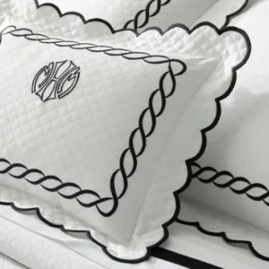 A close-up of a white quilted pillow with black scalloped edges and a monogram in the center, resting on a matching coverlet. Featuring the Classic Chain Scallop Matelassé set.