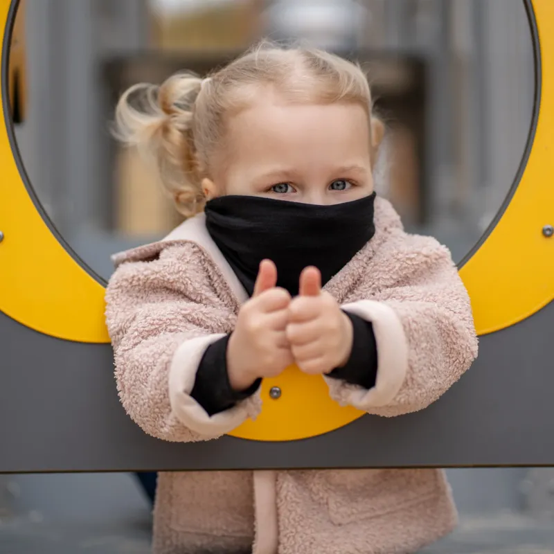 A cheerful child with blonde pigtails looks straight and to the left, peering through a yellow and gray circular opening on a playground. The child is wearing a Black Menique neck gaiter and a fluffy beige jacket, with a slight smile on their face.