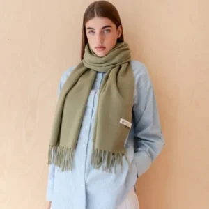 Model wearing a soft Lambswool Oversized Scarf in Olive by TBCo with fringed edges against wooden background