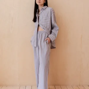 A person wearing TBCo Cotton Trousers in Lilac Stripe with coordinating button-down shirt against wooden background