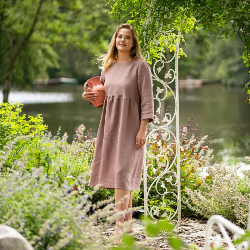 A barefoot woman stands near a white decorative garden archway, wearing a long-sleeve, faded rose linen dress. She smiles softly at the camera while holding a decorative terracotta pitcher comfortably against her side.