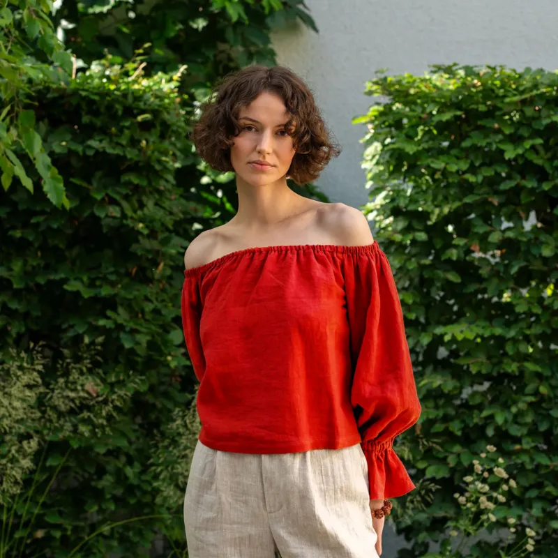 A woman with short, curly brown hair looks directly at the camera, standing in front of a green ivy-covered wall. She is wearing a pure red off-the-shoulder linen top with voluminous sleeves that gather at the wrists, and light-colored pants.