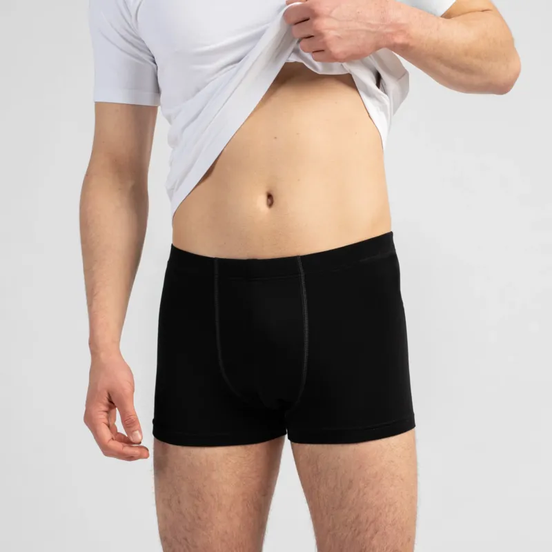 A close-up shot of a man's torso as he pulls up a white t-shirt to reveal his abdomen and black boxer briefs. The background is a plain light grey.