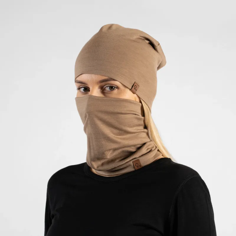 A studio portrait of a blonde woman looking directly at the camera. She is wearing both a light beige slouchy beanie and a matching light beige neck gaiter pulled up to cover her nose and lower face. Both items feature a small brown leather logo patch. She is wearing a solid black long-sleeved top against a plain light grey background.