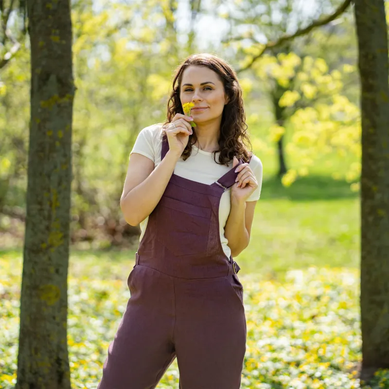 A woman with brown hair, wearing dark purple overalls over a light-colored t-shirt, stands outdoors between two tree trunks on grassy ground.