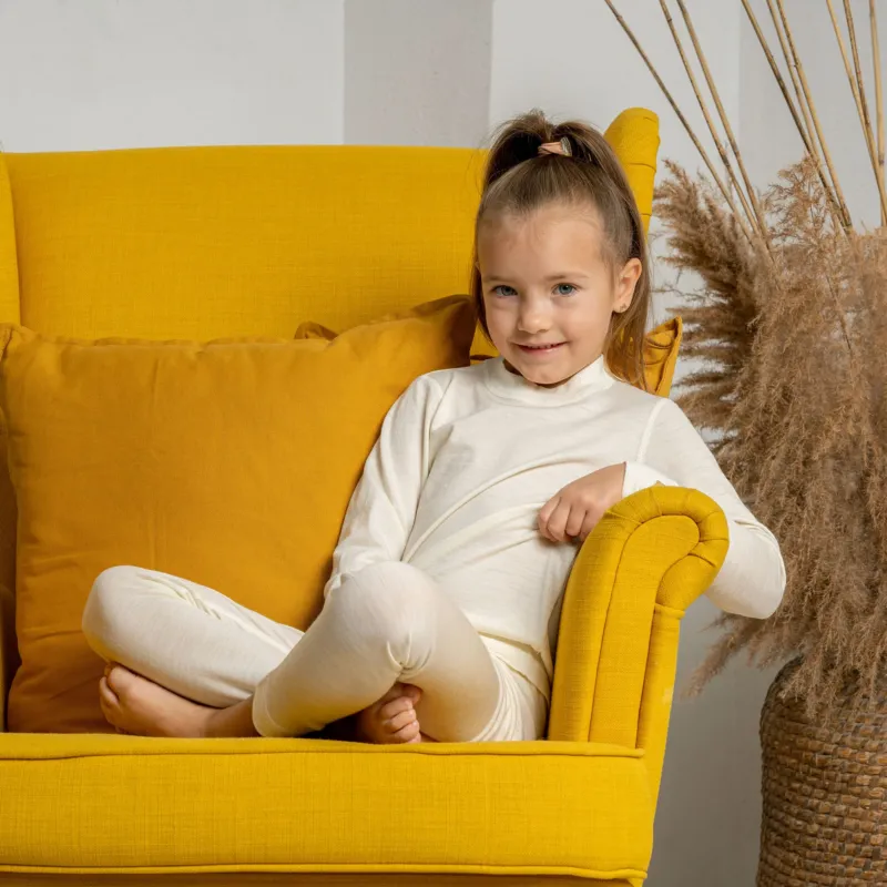 A girl sitting in a yellow armchair, with her legs crossed, wearing the natural color menique long sleeve merino 160gsm top in natural color.