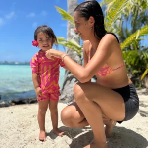 Petite fille portant la combinaison anti-UV ALL-IN Matis Rose sur une plage tropicale avec sa maman