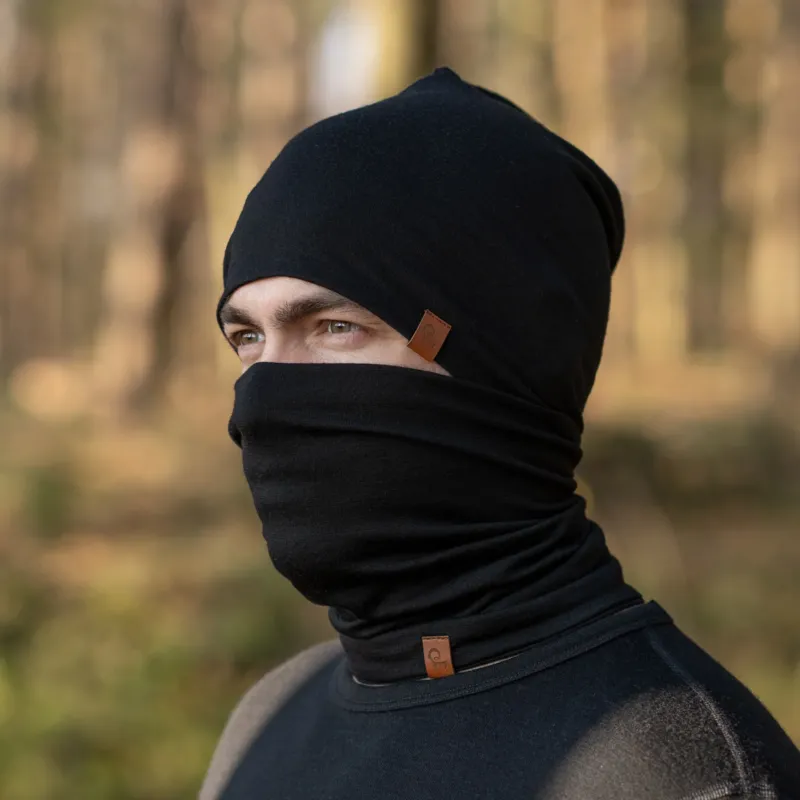 The Menique Men's Merino Beanie and Gaiter Set in Black color. The man is wearing the accessory set in a forest, in fall time.